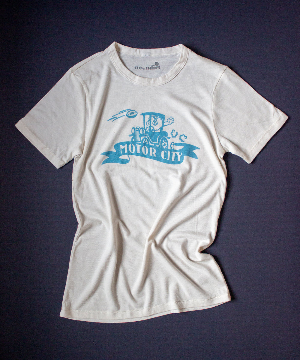 Our Forgotten and Refreshed: Detroit Lions light blue monochrome design features a lion driving a Model T automobile centered on the tee. A football soars across the shirt from the right shoulder. On the lower front center of the design features a banner with text that reads "motor city".