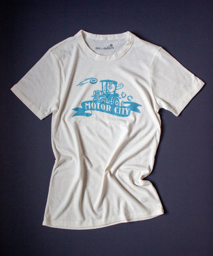 Our Forgotten and Refreshed: Detroit Lions light blue monochrome design features a lion driving a Model T automobile centered on the tee. A football soars across the shirt from the right shoulder. On the lower front center of the design features a banner with text that reads "motor city".