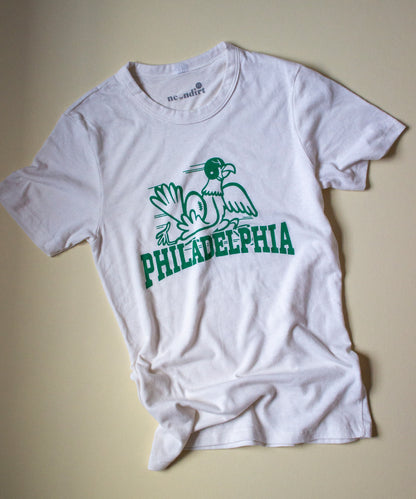 Our Vintage White Forgotten and Refreshed: Philadelphia Eagles tee highlights a green monochrome design which features an eagle wearing a cracked helmet, rushing towards the left shoulder. In the foreground vintage inspired text reading "Philadelphia" covers the lower half of the design, horizontally. 